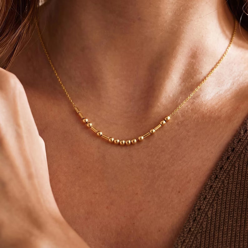 Close-up of a gold tone beaded Morse code necklace resting on the collarbone above a brown knit top