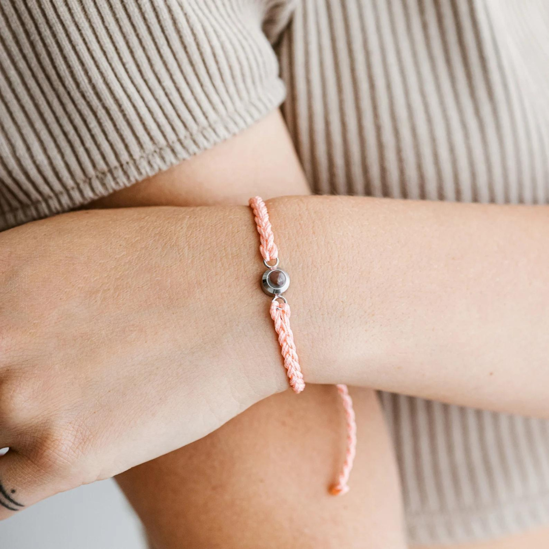 Peach pink braided cord bracelet with a small silver photo stone worn on a woman's wrist over a ribbed top
