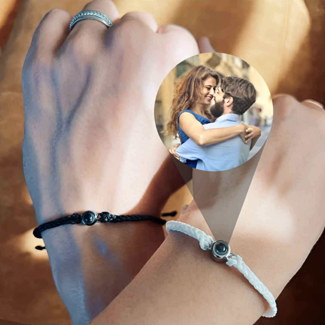 Two hands wearing black and white cord photo bracelets, the stone projecting a photo of a hugging couple