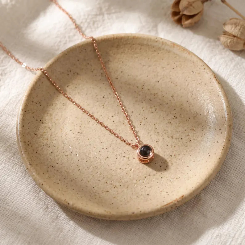 Rose gold custom photo necklace with a tiny round pendant laid on a speckled ceramic dish on linen